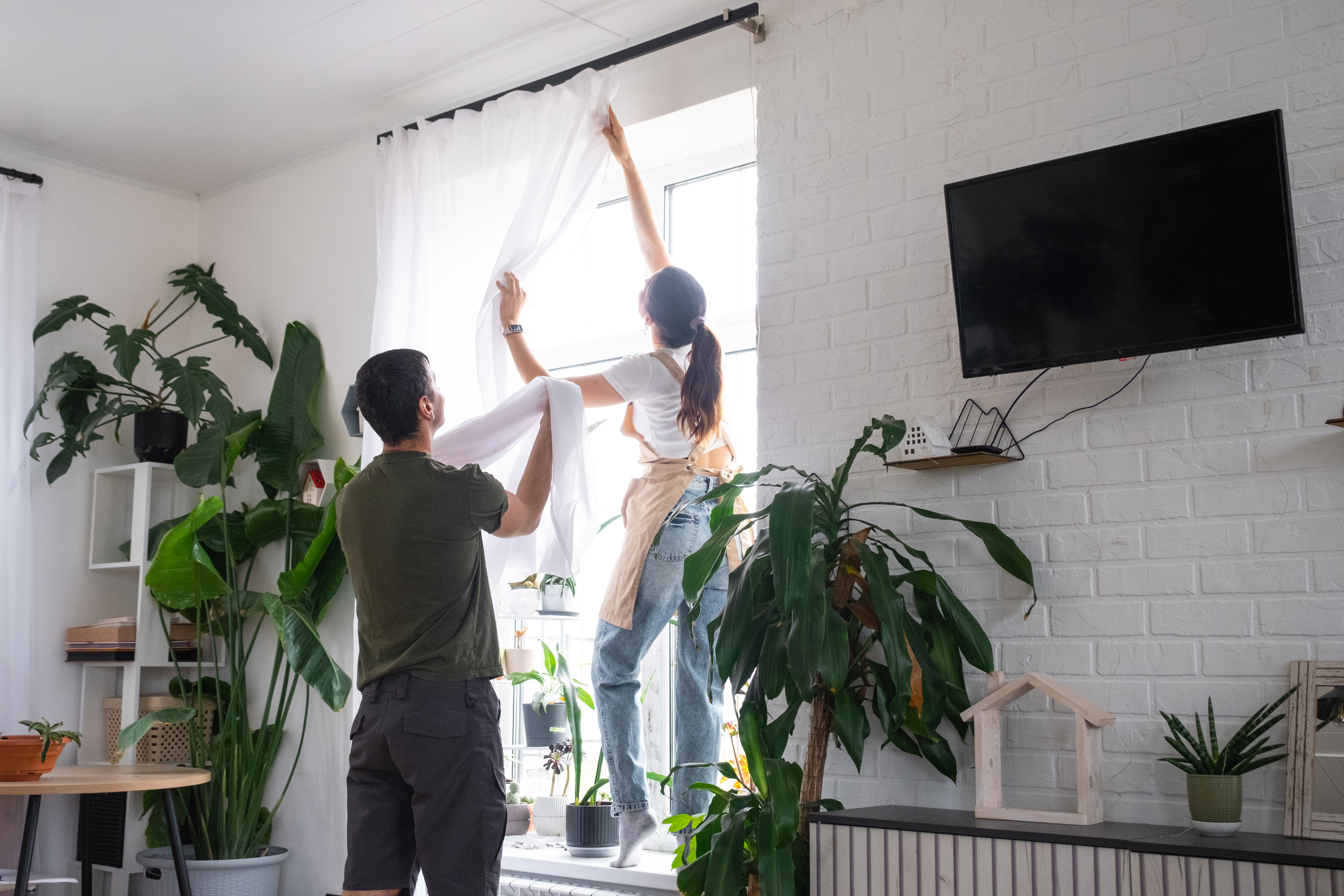 Two people opening curtains to let in fresh air and sunlight while cleaning a bright, plant-filled home.